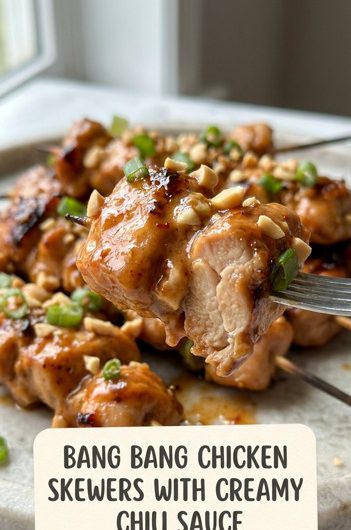 Golden bang bang chicken skewers on a white plate, drizzled with creamy orange chili sauce and garnished with sliced green onions